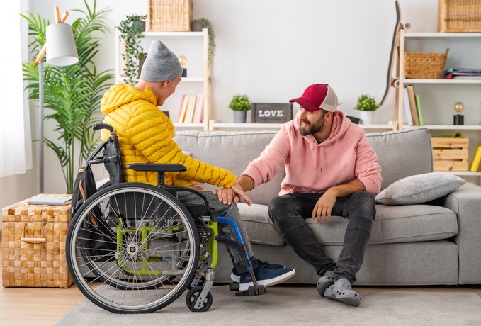 Zwei Männer sitzen im Wohnzimmer auf einem Sofa, einer von ihnen im Rollstuhl